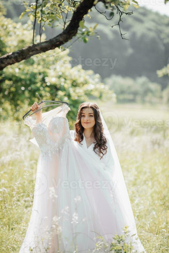 lindo noiva com grandes encaracolado cabelo dentro uma verde jardim, segurando dela delicado vestir, olhando para dentro a lente. manhã do a noiva. natural inventar. Boa luz do dia. foto