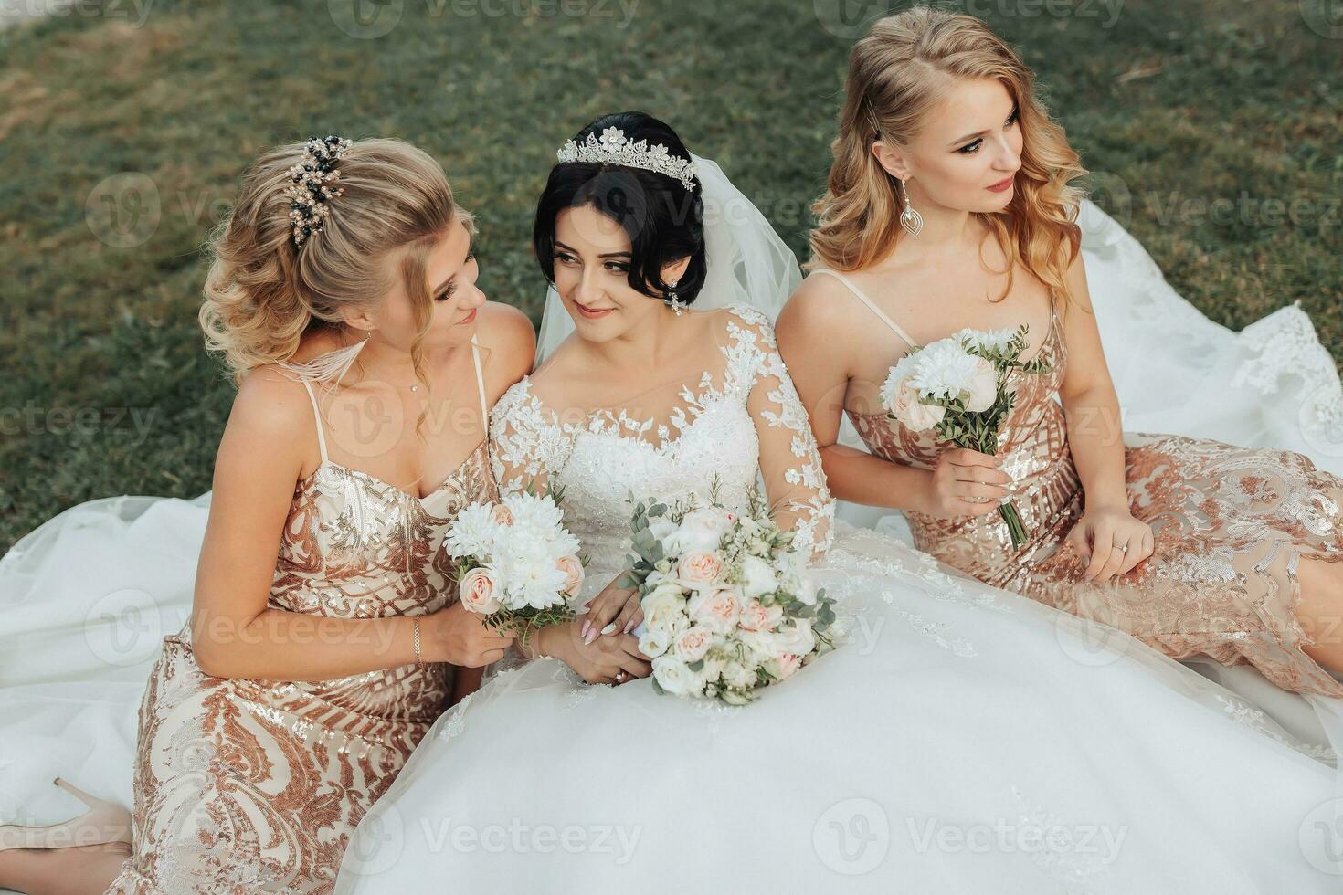 uma morena noiva dentro uma branco elegante vestir com uma coroa e dela Loiras amigos dentro ouro vestidos pose com buquês enquanto sentado em a grama. Casamento retrato dentro natureza, Casamento foto dentro uma luz tom.