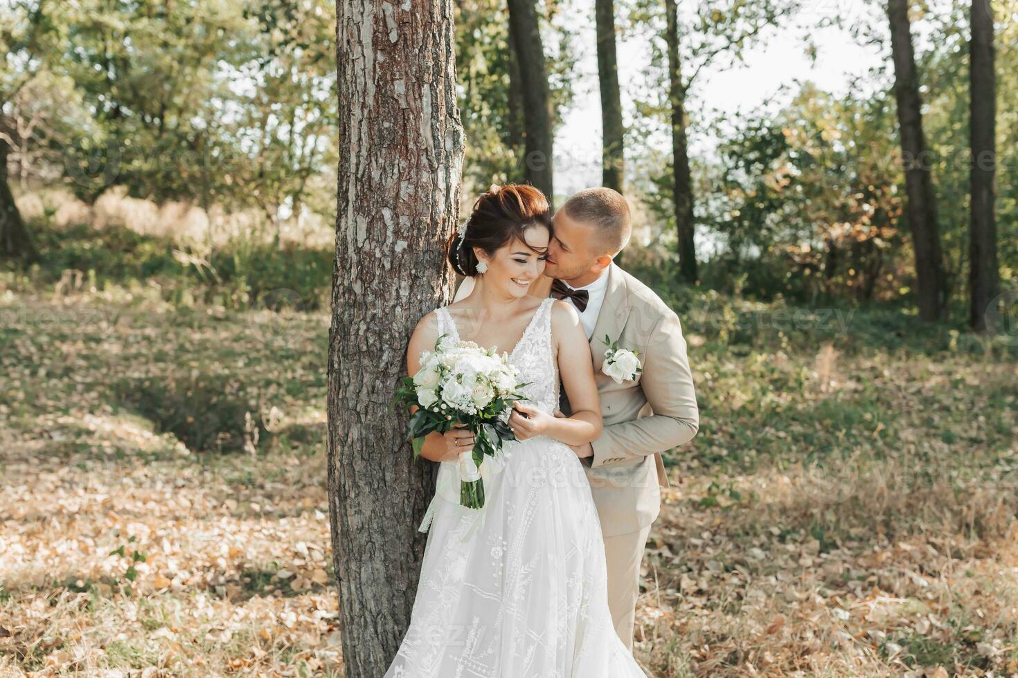 Casamento foto dentro natureza. a noiva e noivo estão em pé perto uma árvore, a noivo abraços dele Amado a partir de atrás e Beijos dela, ela sorrisos sinceramente. retrato. verão Casamento