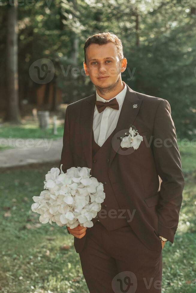 jovem elegante noivo dentro a floresta com uma ramalhete do branco flores dentro uma Castanho terno e uma arco gravata foto
