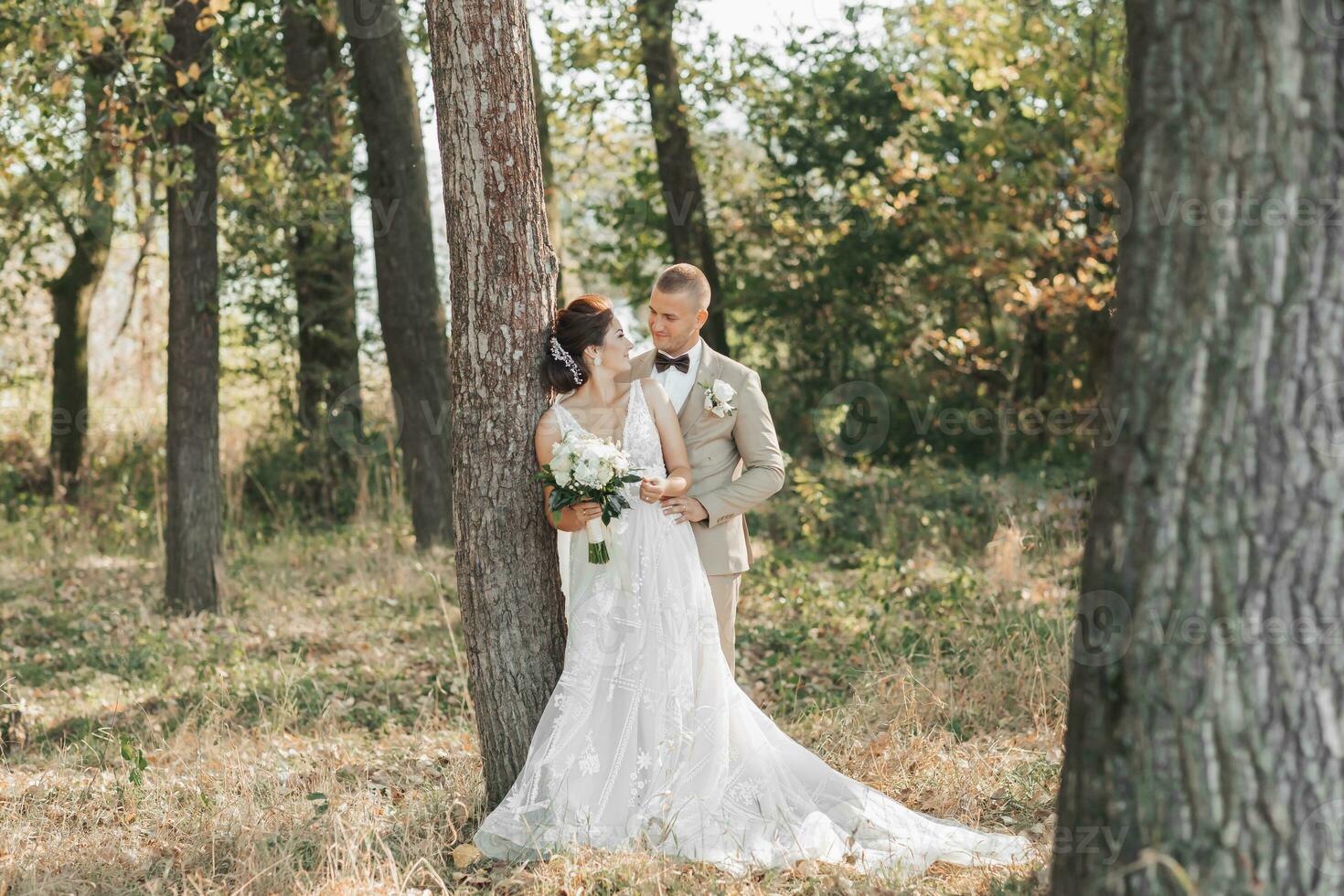 Casamento foto dentro natureza. a noiva e noivo estão em pé perto uma árvore sorridente e olhando às cada outro. a noivo abraços dele Amado a partir de atrás, a noiva detém uma ramalhete. retrato. verão Casamento