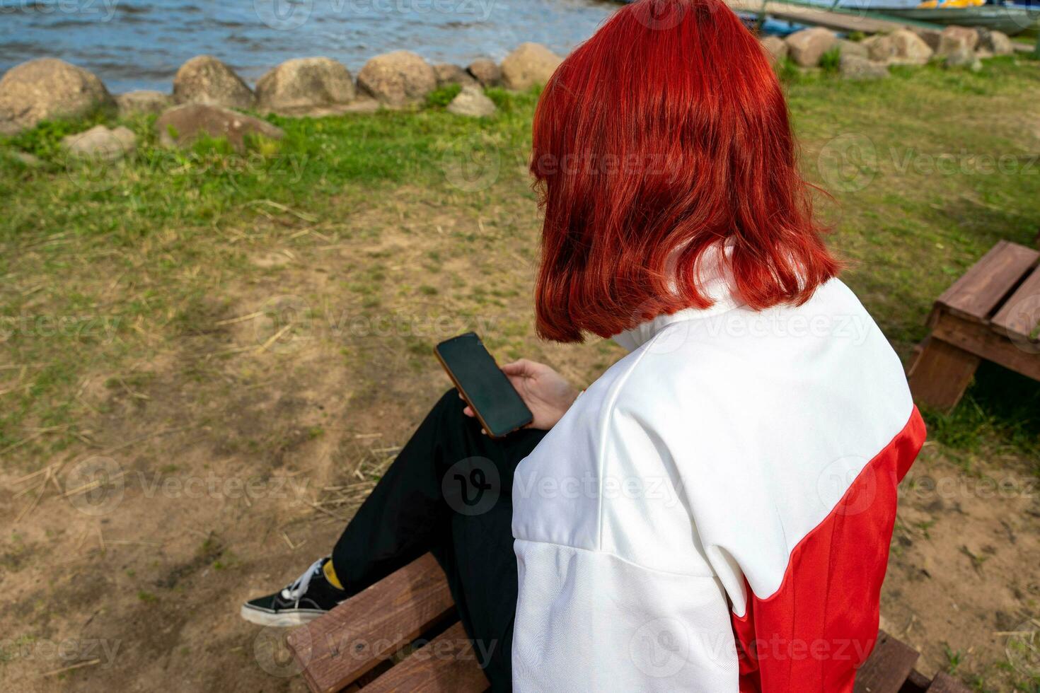 menina com vermelho cabelo, geração z, senta em a costa do uma quieto lago e usa uma Smartphone foto