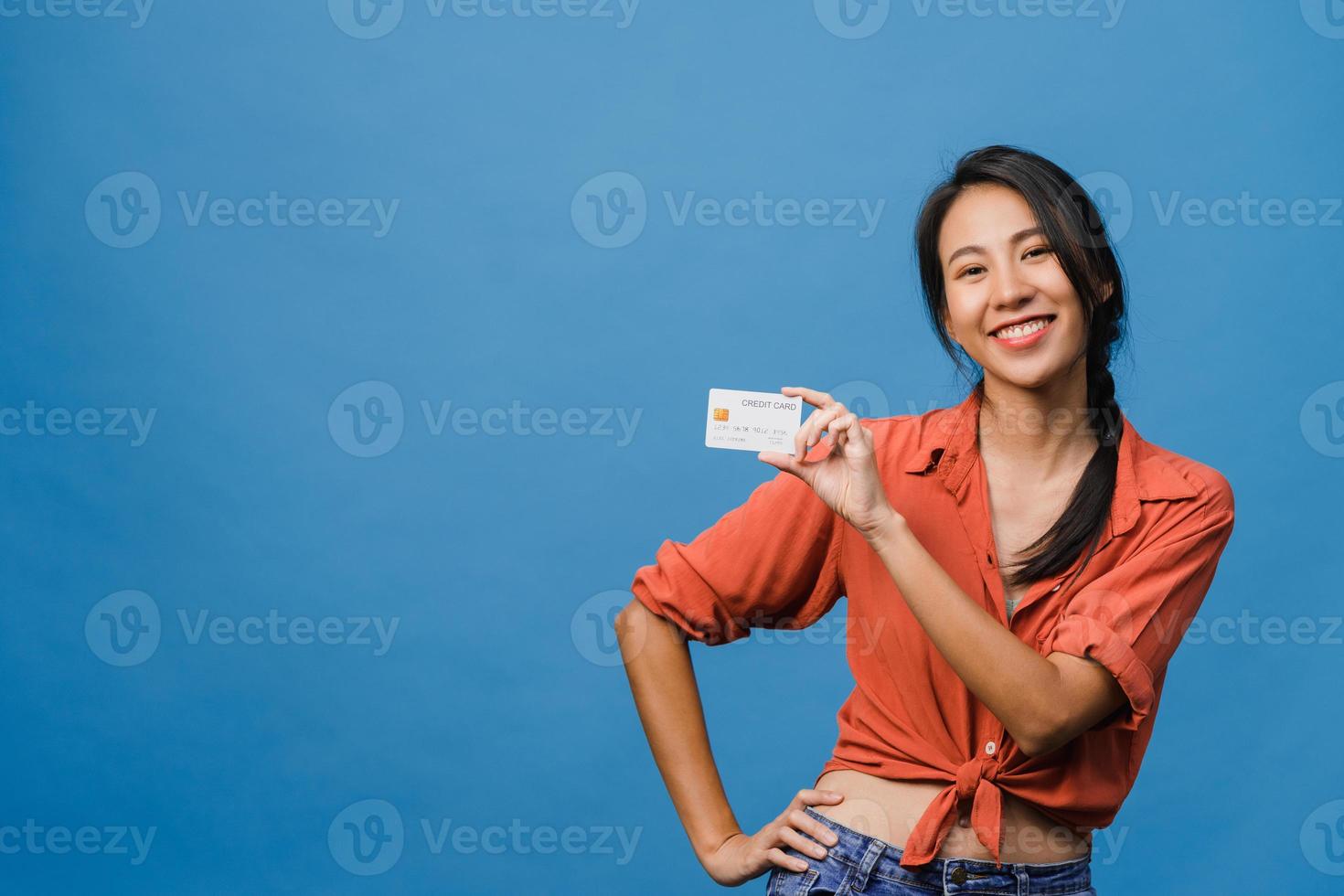 jovem senhora asiática mostra cartão de crédito com expressão positiva, sorri amplamente, vestida com roupas casuais, sentindo felicidade e carrinho isolado sobre fundo azul. conceito de expressão facial. foto