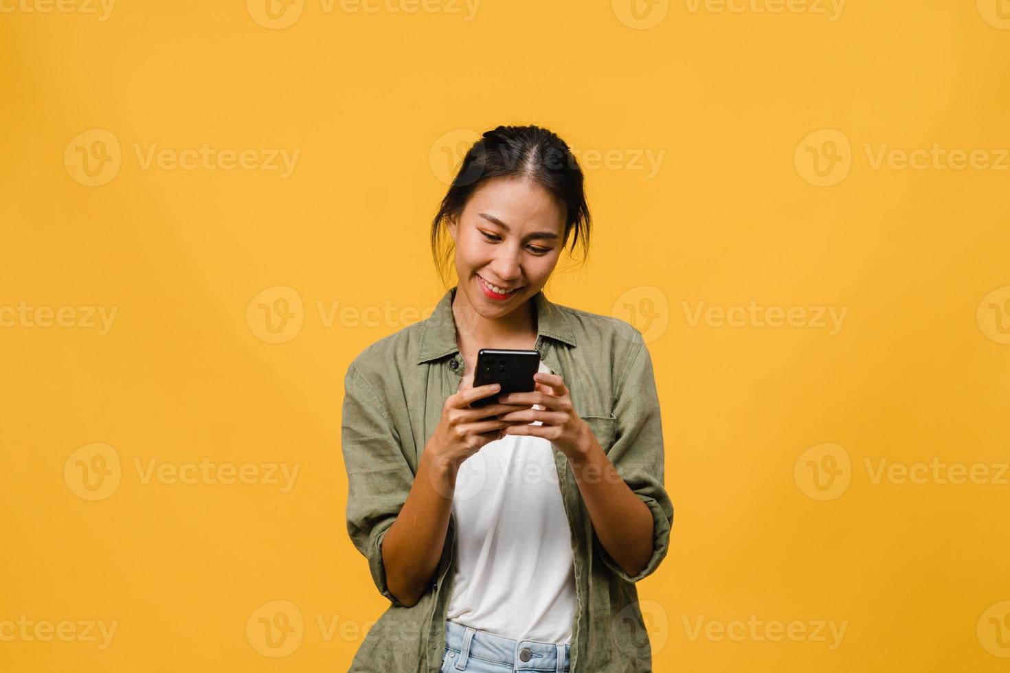 jovem asiática usando telefone com expressão positiva, sorri amplamente, vestida com roupas casuais, sentindo felicidade e carrinho isolado em fundo amarelo. feliz adorável feliz mulher alegra sucesso. foto
