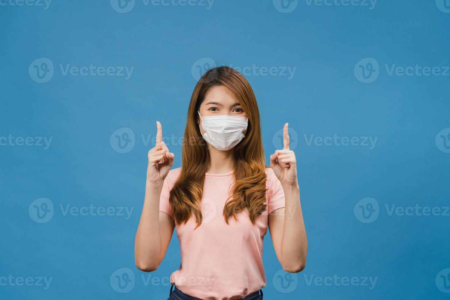 jovem asiática usando máscara médica mostra algo no espaço em branco com vestido de pano casual e olhando para a câmera isolada sobre fundo azul. distanciamento social, quarentena para o vírus corona. foto