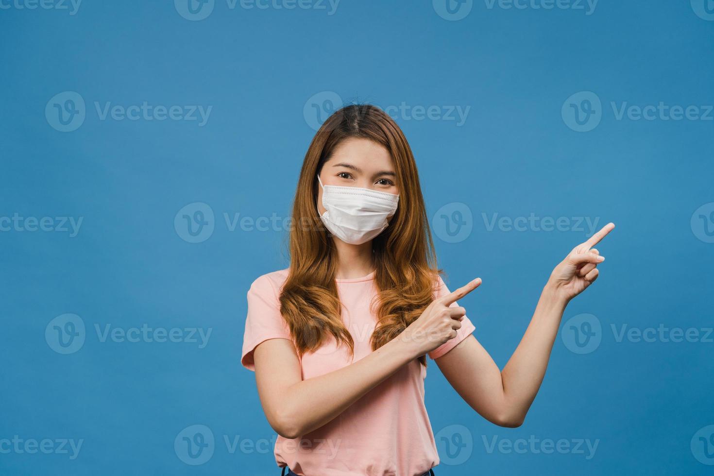 jovem asiática usando máscara médica mostra algo no espaço em branco com vestido de pano casual e olhando para a câmera isolada sobre fundo azul. distanciamento social, quarentena para o vírus corona. foto