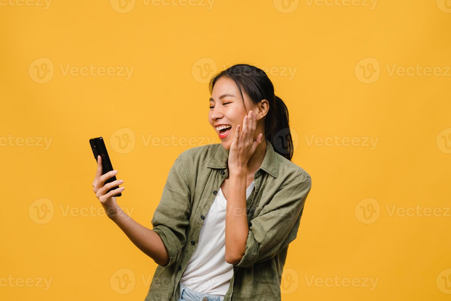 jovem asiática usando telefone com expressão positiva, sorri amplamente, vestida com roupas casuais, sentindo felicidade e carrinho isolado em fundo amarelo. feliz adorável feliz mulher alegra sucesso. foto