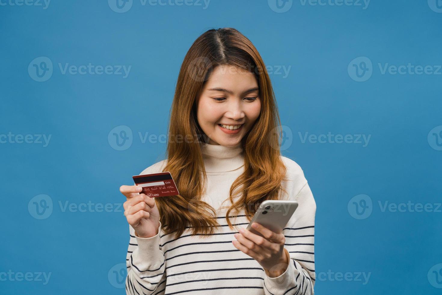 jovem asiática usando telefone e cartão de crédito com expressão positiva, sorri amplamente, vestida com roupas casuais e carrinho isolado sobre fundo azul. feliz adorável feliz mulher alegra sucesso. foto