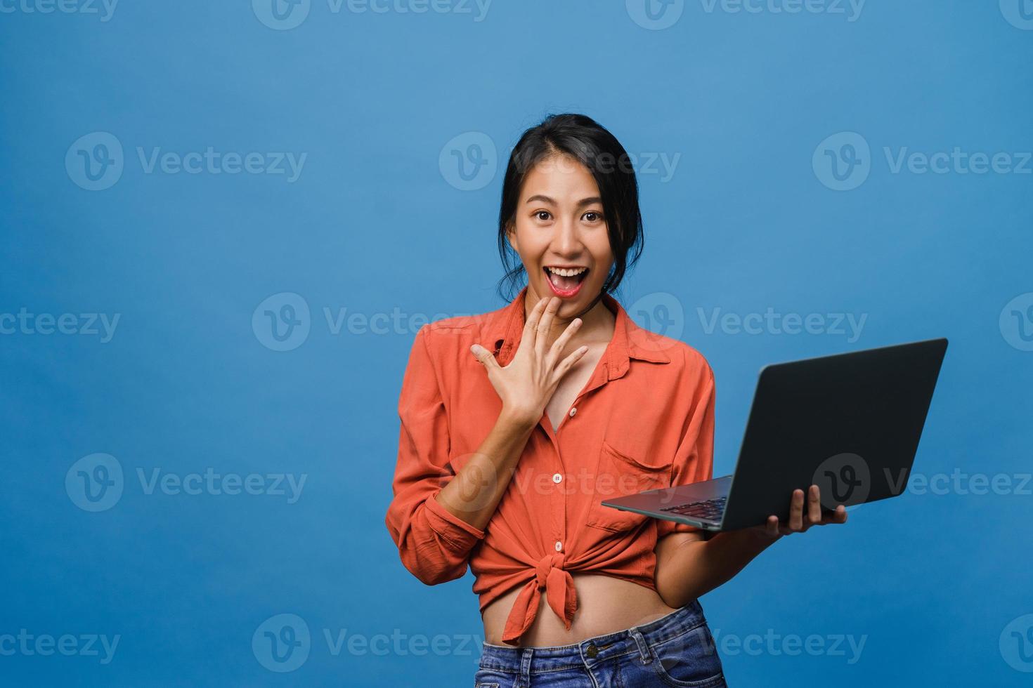 surpreendeu a jovem asiática usando laptop com expressão positiva, sorriso largo, vestido com roupas casuais e olhando para a câmera sobre fundo azul. feliz adorável feliz mulher alegra sucesso. foto