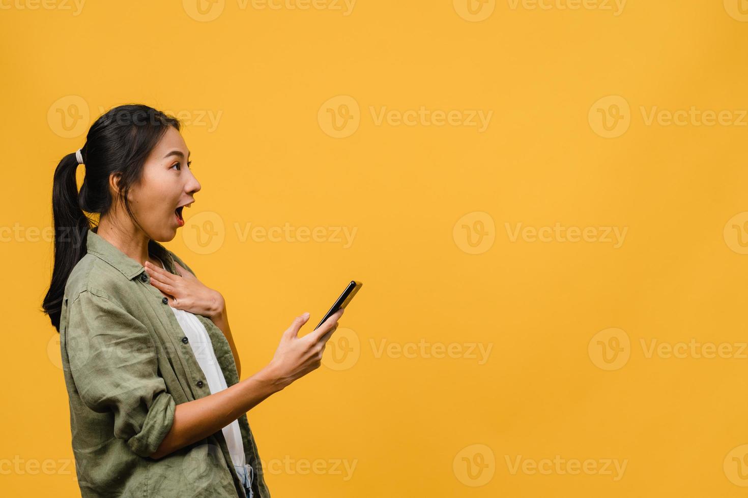 surpreendeu a jovem asiática usando telefone celular com expressão positiva, sorri amplamente, vestida com roupas casuais e carrinho isolado em fundo amarelo. feliz adorável feliz mulher alegra sucesso. foto