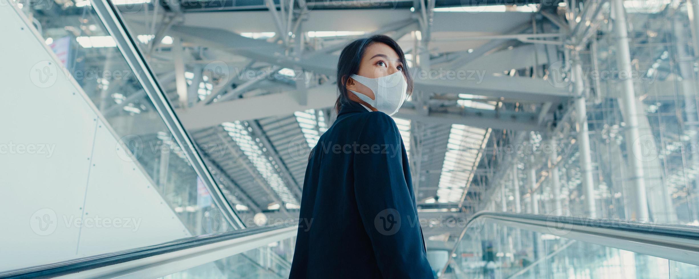 garota de negócios asiáticos usar máscara facial arraste carrinho de bagagem na escada rolante olhar ao redor caminhar para o terminal no aeroporto internacional. conceito de distanciamento social de viagens de negócios. fundo de banner panorâmico. foto