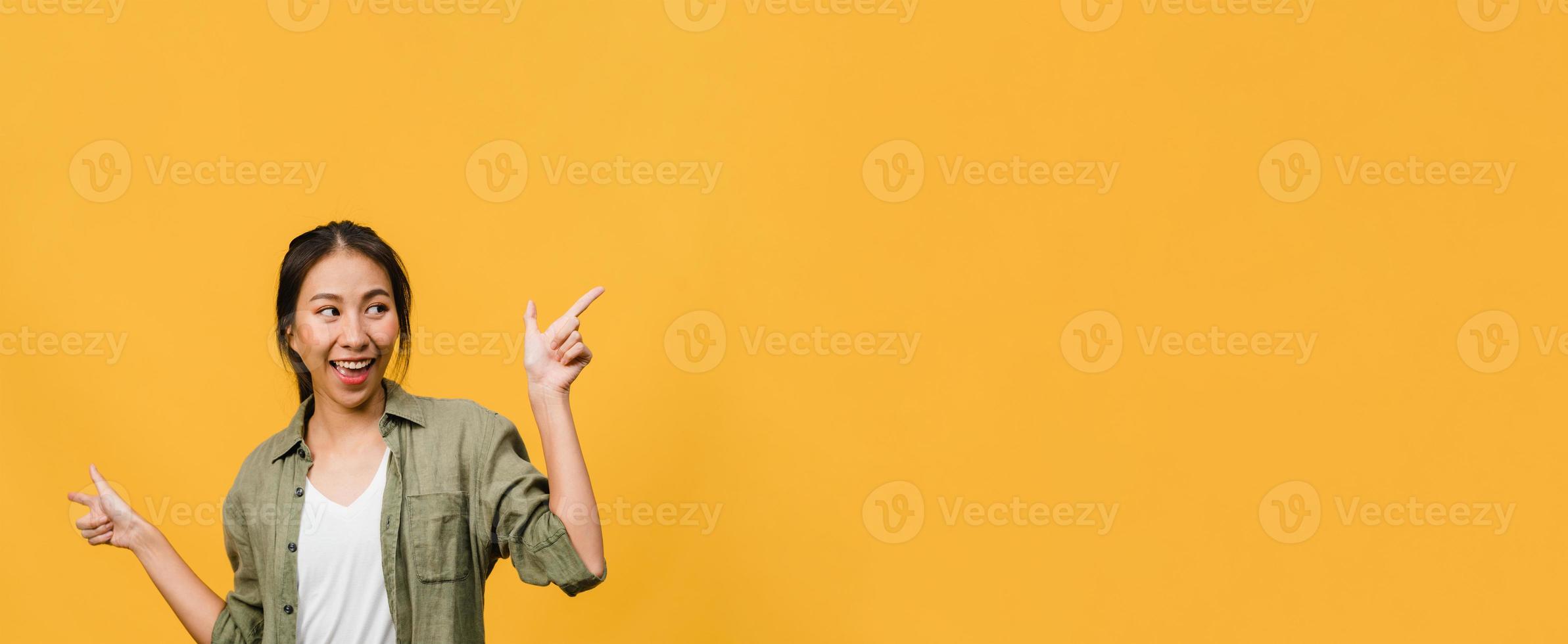 retrato de uma jovem senhora asiática, sorrindo com uma expressão alegre, mostra algo incrível no espaço em branco em roupas casuais e em pé isolado sobre fundo amarelo. banner panorâmico com espaço de cópia. foto