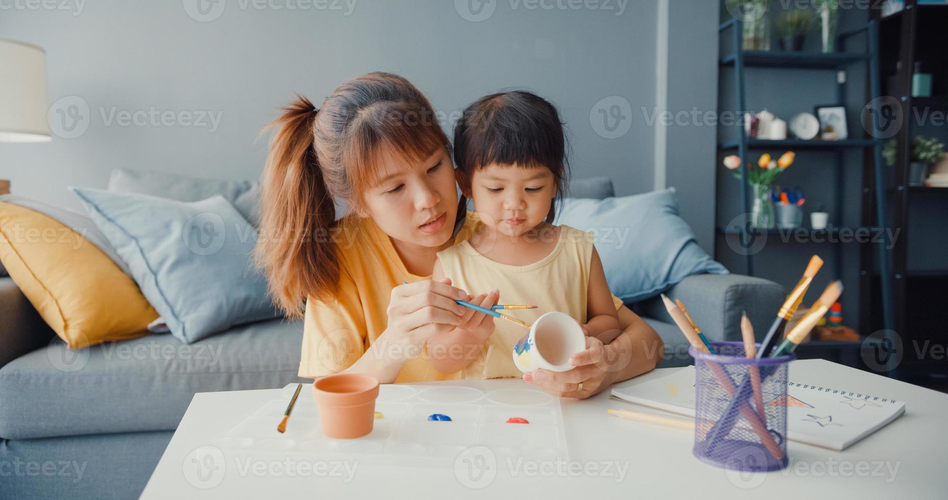 feliz alegre mãe de família asiática ensinar menina criança pintar pote de cerâmica se divertindo, relaxar na mesa na sala de estar em casa. passar tempo juntos, distância social, quarentena para prevenção do coronavírus. foto