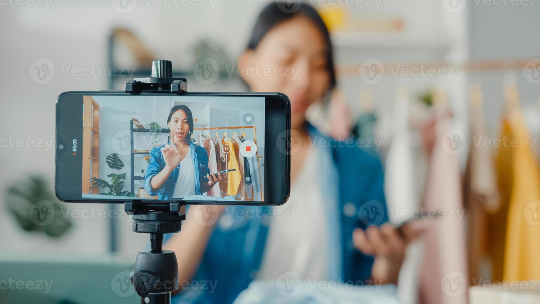 designer de moda jovem asiática usando telefone celular, recebendo pedido de compra e mostrando roupas, gravando vídeo com transmissão ao vivo online na loja. proprietário de uma pequena empresa, conceito de entrega de mercado online. foto