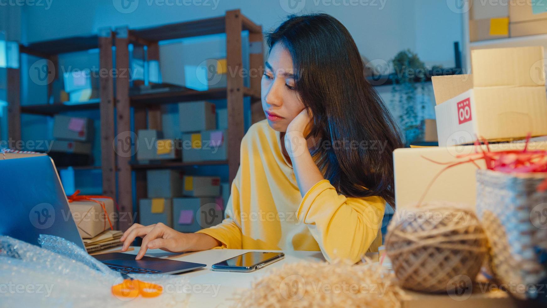 jovem empresária da ásia olha ao redor uma sala cheia de coisas de produtos e caixa de encomendas sente estresse e chateada com vendas ruins no escritório em casa à noite. proprietário de uma pequena empresa, conceito de entrega de mercado online. foto