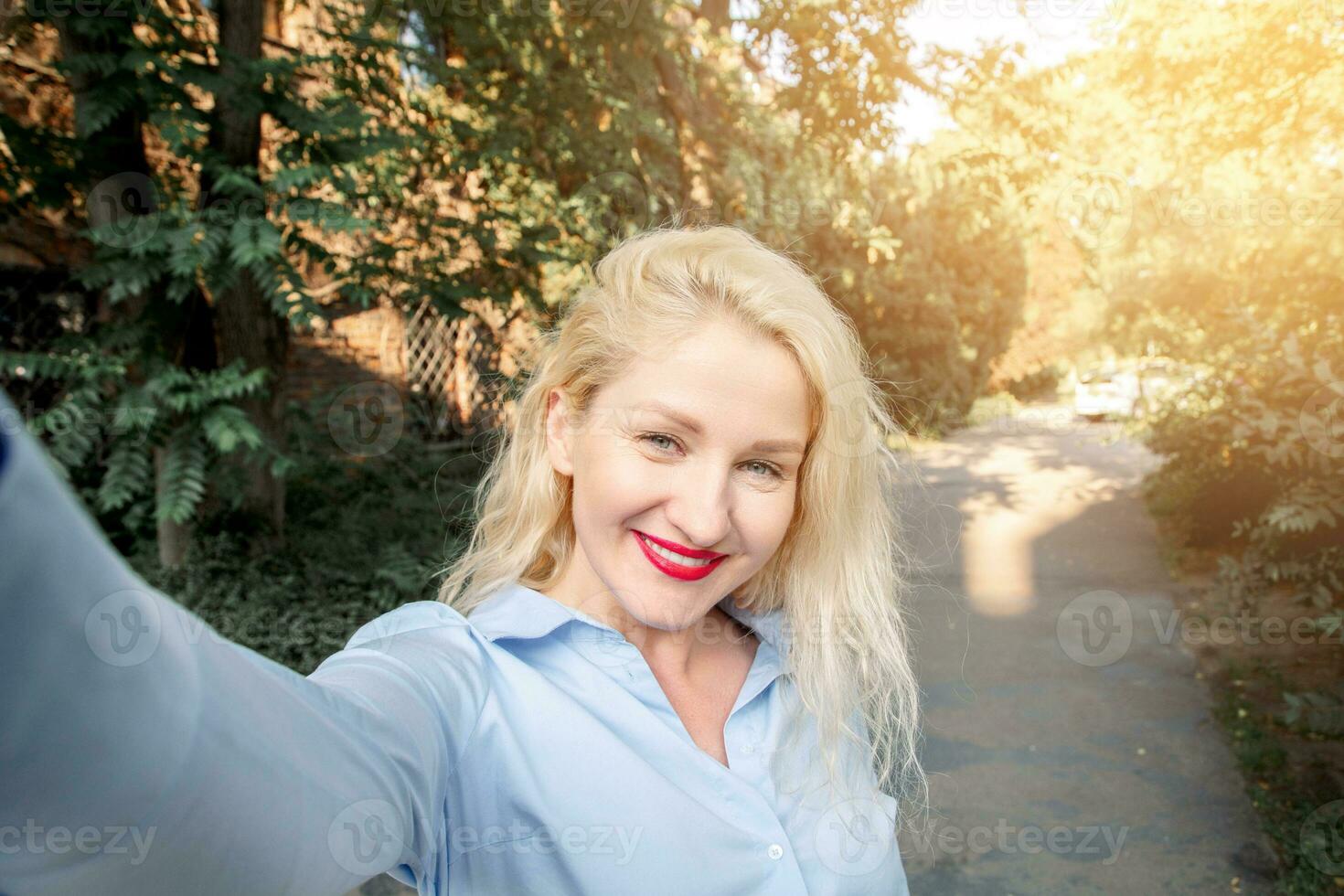 feliz menina com Loiras cabelo vestido dentro vestindo uma azul camisa, caminhando por aí a cidade, posando para selfies. uma caloroso verão tarde. foto