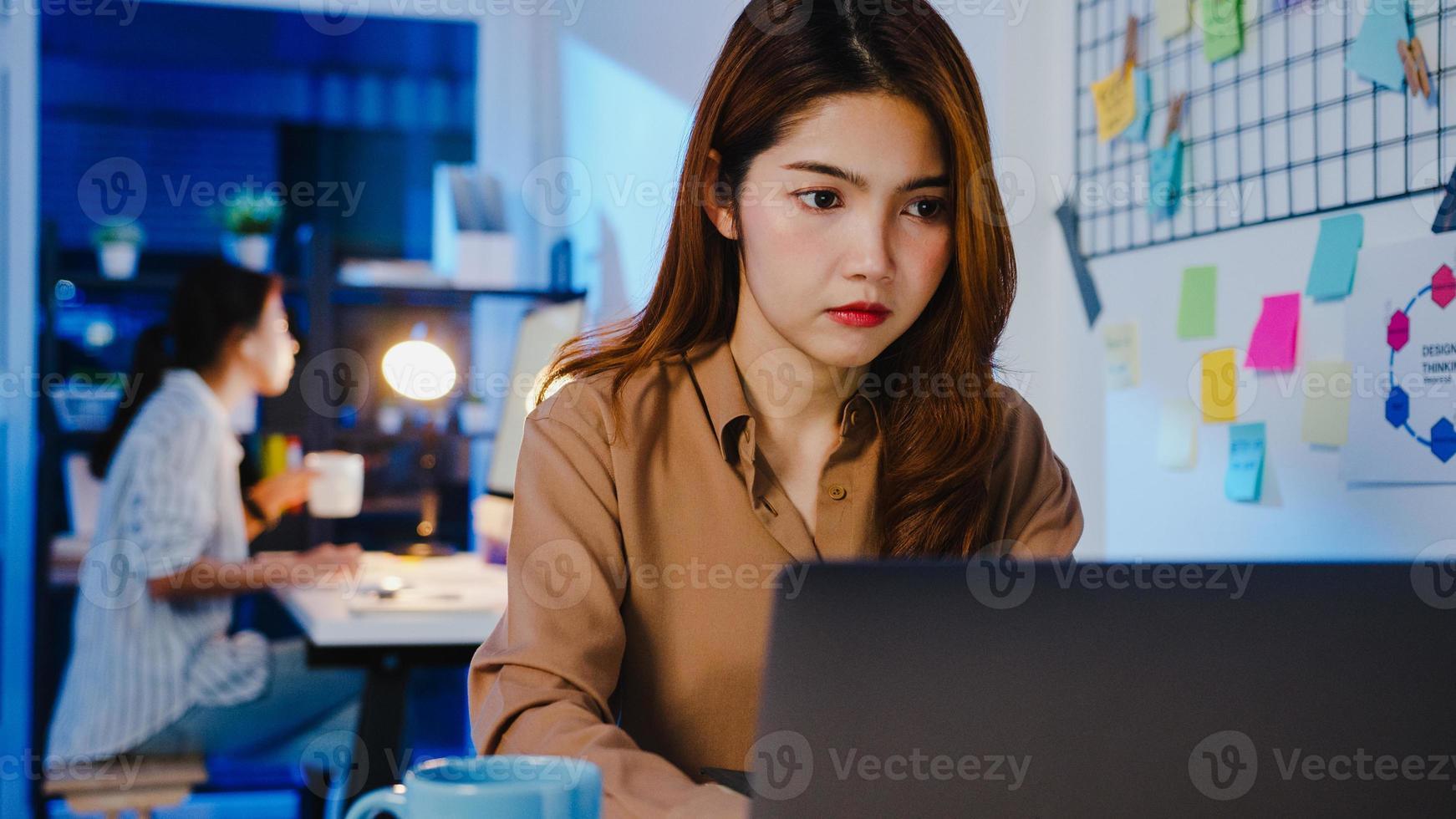 Feliz Ásia empresária distanciamento social na nova situação normal para prevenção de vírus ao usar o laptop de negócios on-line horas extras no trabalho, à noite no escritório. vida e trabalho após o coronavírus. foto
