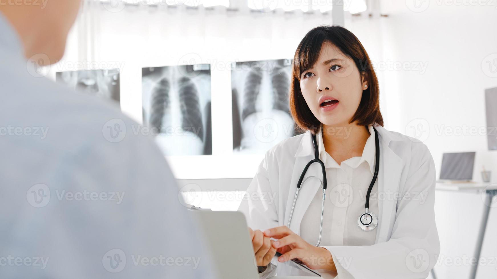 jovem médica asiática em uniforme médico branco, usando o computador laptop, está dando uma ótima palestra, discute os resultados ou os sintomas com um paciente do sexo masculino sentado na mesa na clínica de saúde ou no escritório do hospital. foto