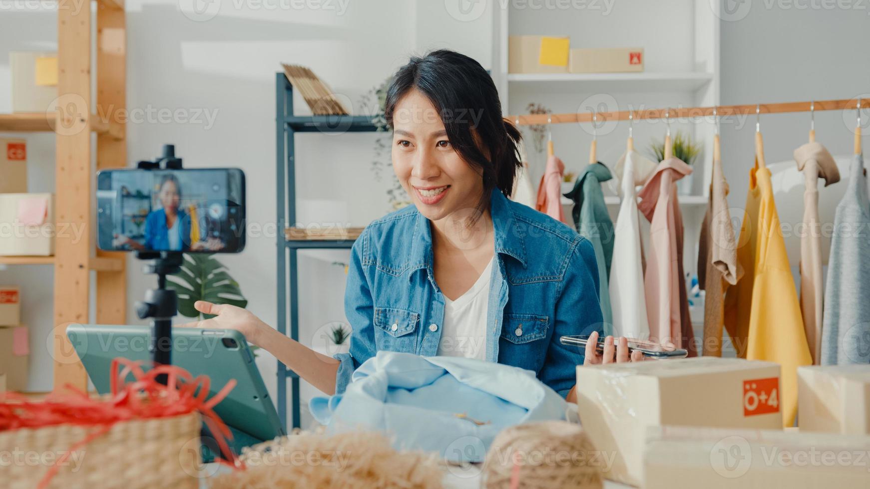 designer de moda jovem asiática usando telefone celular, recebendo pedido de compra e mostrando roupas, gravando vídeo com transmissão ao vivo online na loja. proprietário de uma pequena empresa, conceito de entrega de mercado online. foto