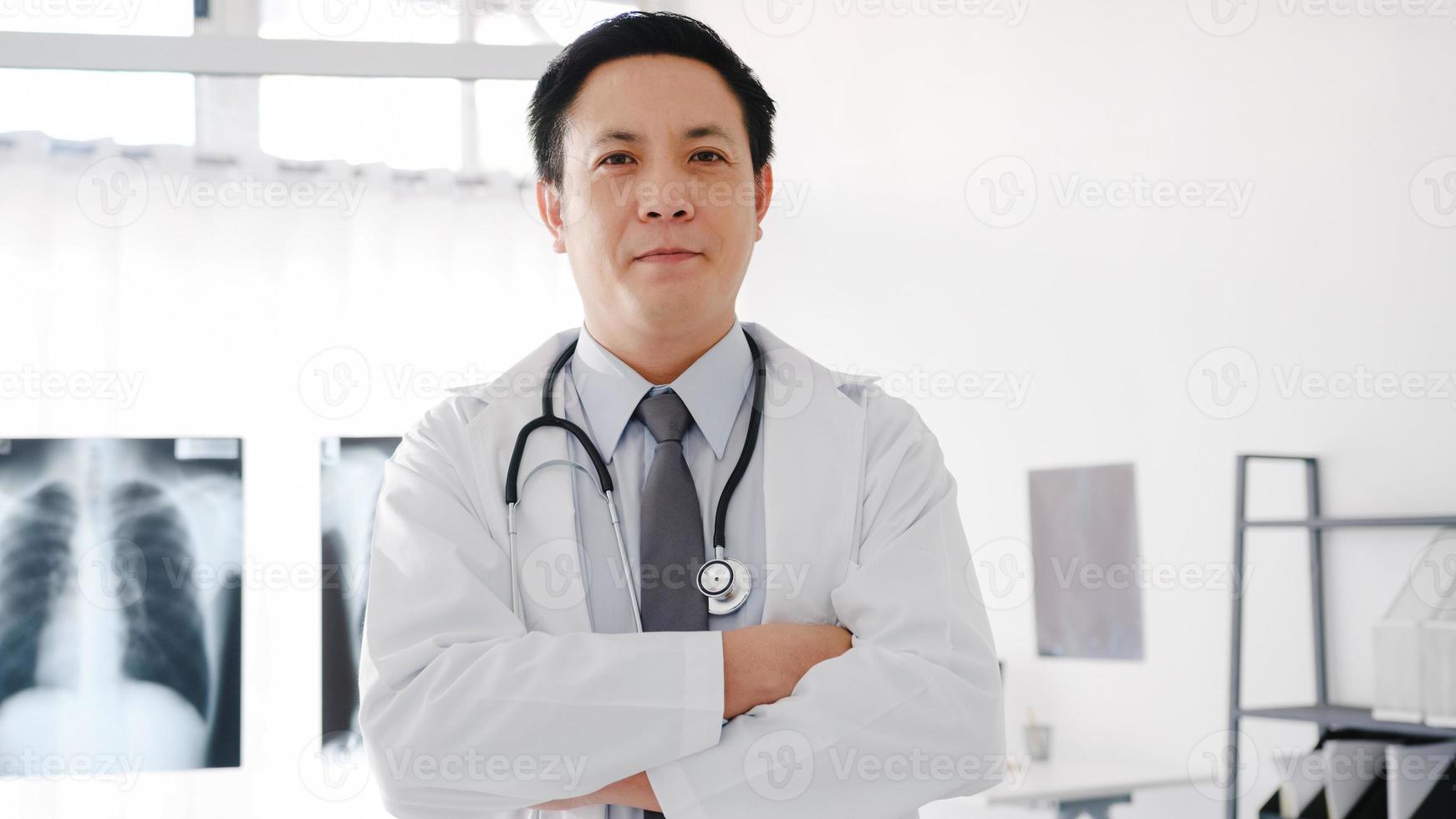 jovem médico asiático em uniforme médico branco com estetoscópio olhando para a câmera, sorriso e braços cruzados durante a videoconferência com o paciente no hospital de saúde. consultoria e conceito de terapia. foto