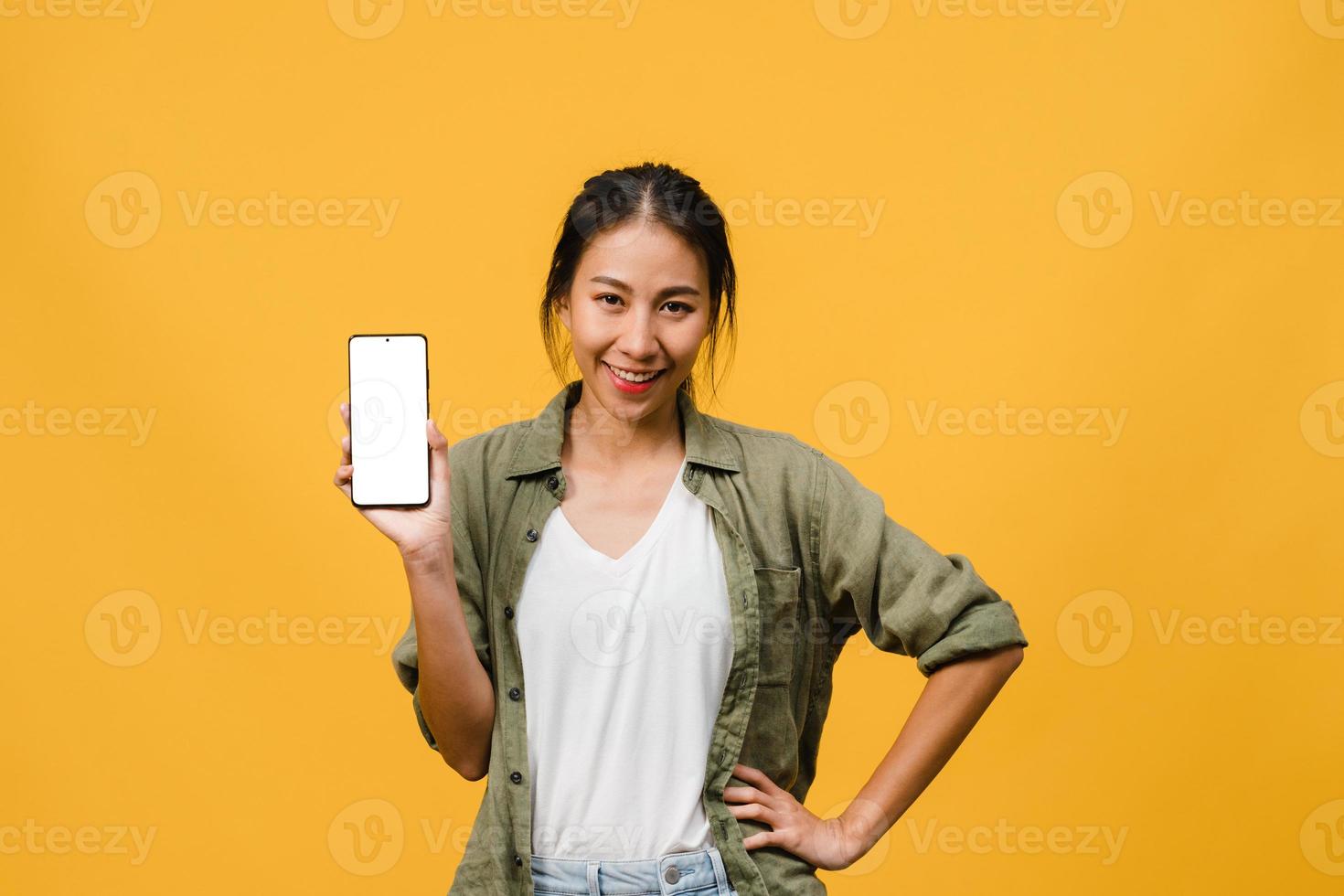 jovem senhora asiática mostra tela vazia do smartphone com expressão positiva, sorri amplamente, vestida com roupas casuais, sentindo felicidade sobre fundo amarelo. telefone celular com tela branca na mão feminina. foto