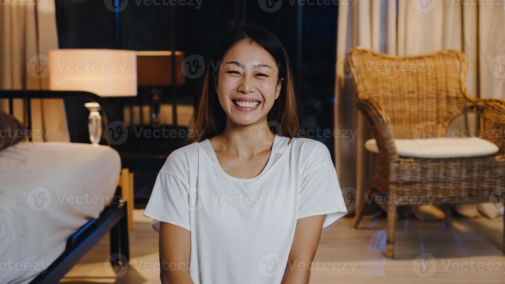 alegre jovem asiática se sentindo feliz sorrir e olhar para a câmera usando o telefone para fazer videochamada ao vivo na sala de estar em casa à noite. distanciamento social, quarentena para coronavírus. close-up visualização da webcam. foto
