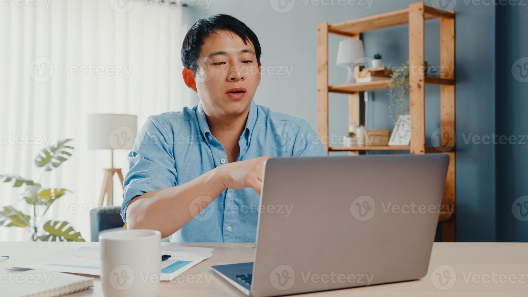 jovem empresário asiático usando laptop fala com colegas sobre o plano de videochamada enquanto inteligente trabalhando em casa na sala de estar. auto-isolamento, distanciamento social, quarentena para prevenção do vírus corona. foto