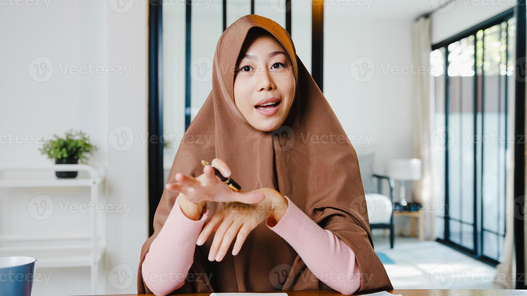 A senhora muçulmana asiática usa o hijab usando um laptop para conversar com colegas sobre o plano de uma reunião de videochamada enquanto trabalha remotamente em casa na sala de estar. distanciamento social, quarentena para o vírus corona. foto