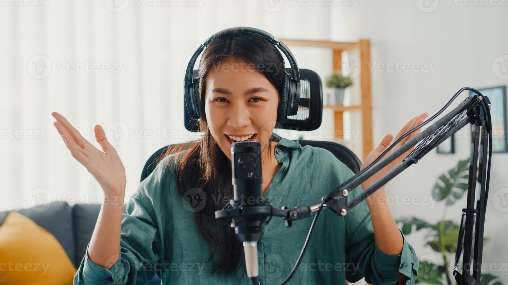 menina asiática feliz gravar um podcast com fones de ouvido e microfone olhar para a câmera falar e descansar no quarto dela. podcaster feminino fazer podcast de áudio de seu estúdio em casa, conceito de ficar em casa. foto