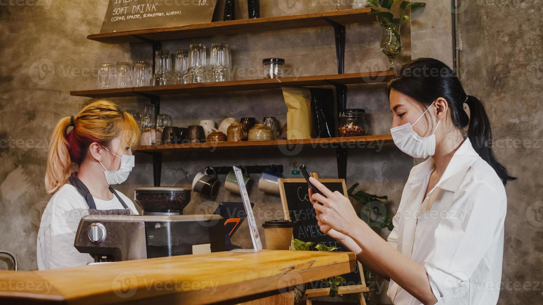 jovem ásia feminina usar máscara facial self-service usar telefone celular pagar sem contato no restaurante. menina barista falando receber o pedido do cliente no café. estilo de vida novo normal após o conceito de vírus corona. foto