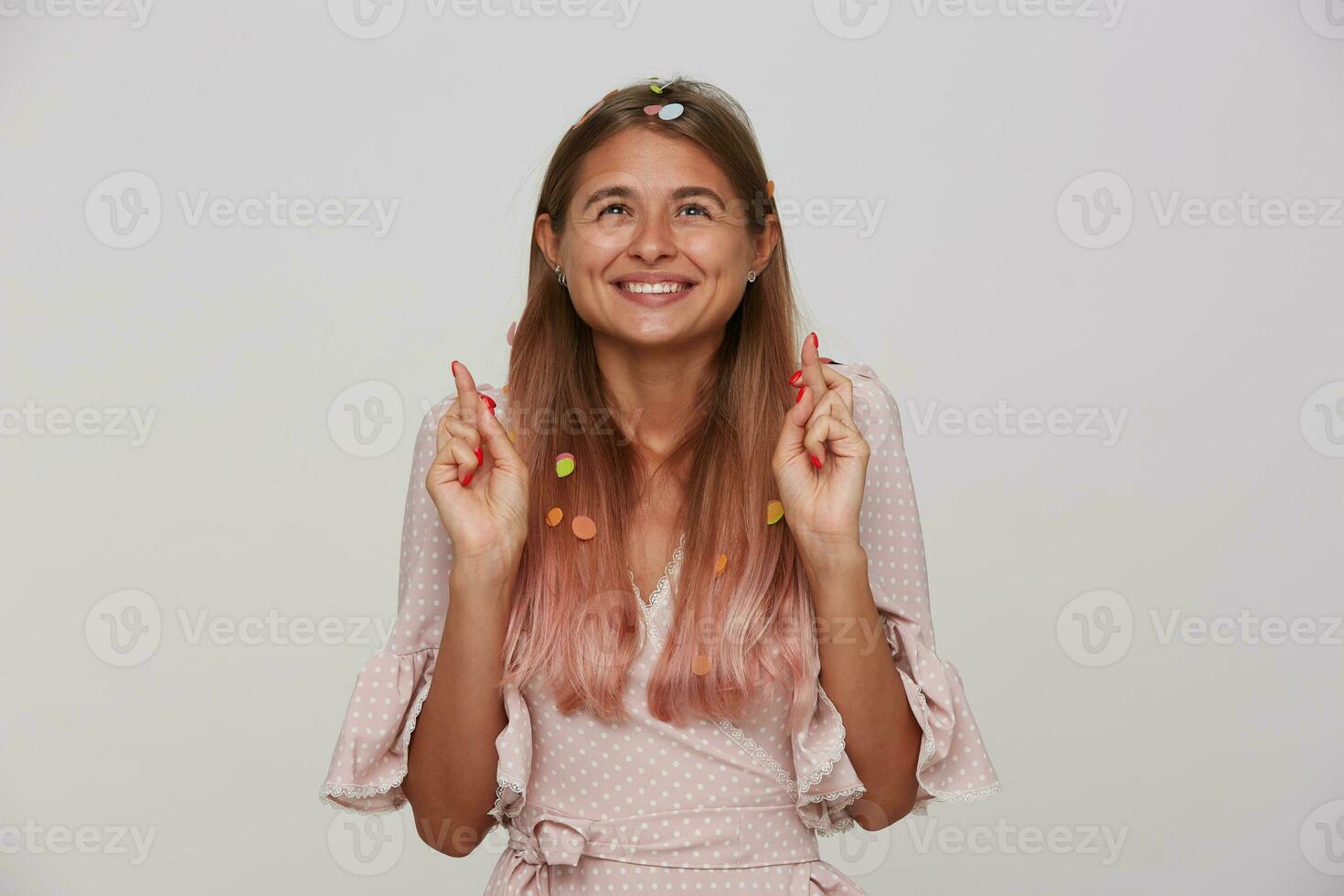 estúdio foto do alegre jovem lindo Loiras senhora dentro festivo elegante roupas sorridente positivamente e levantando mãos com cruzado dedos enquanto em pé sobre branco fundo