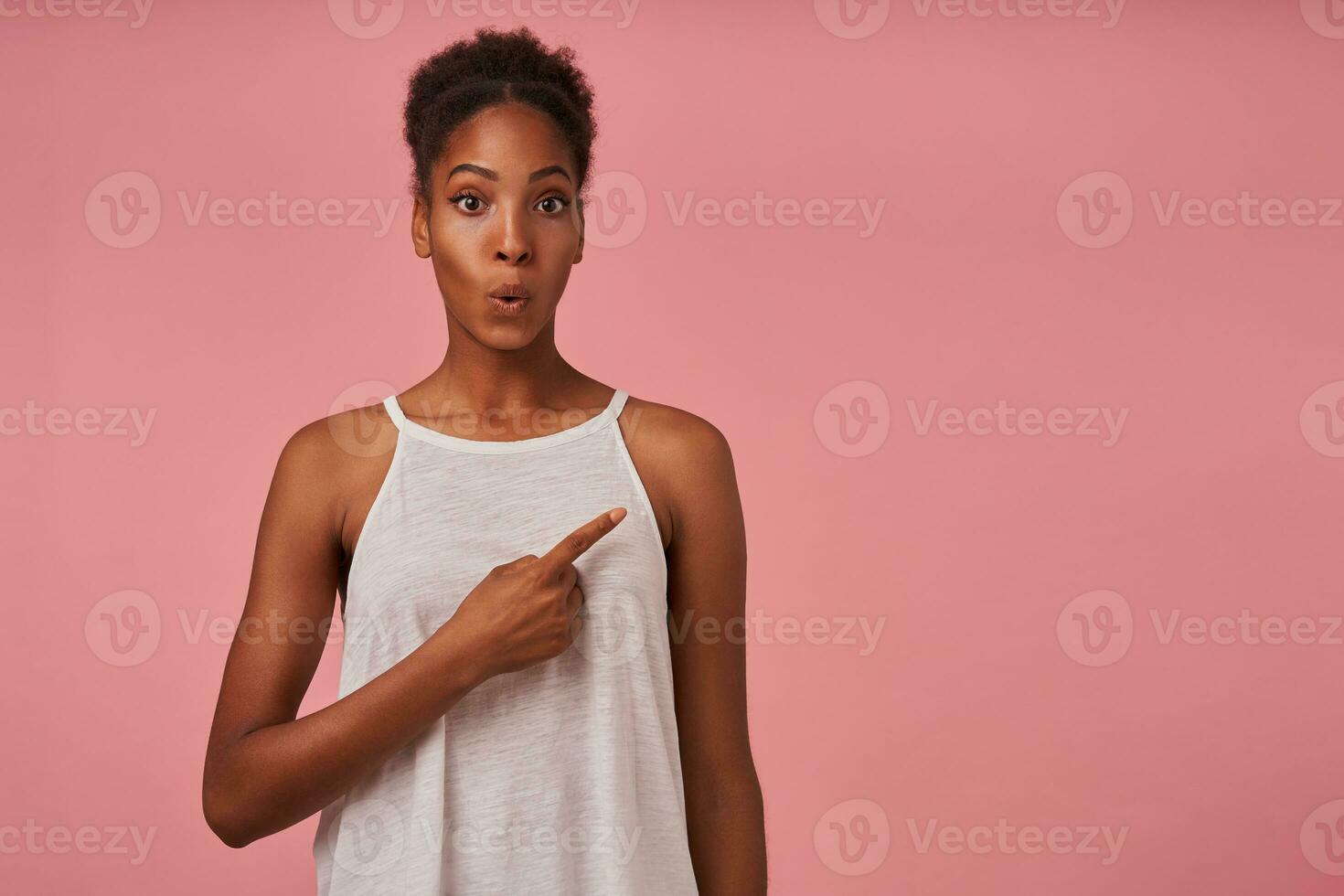 agitado jovem lindo Castanho cabelos Sombrio esfolado senhora levantando surpreendentemente dela sobrancelhas enquanto apontando a parte, de lado com índice dedo, em pé sobre Rosa fundo foto