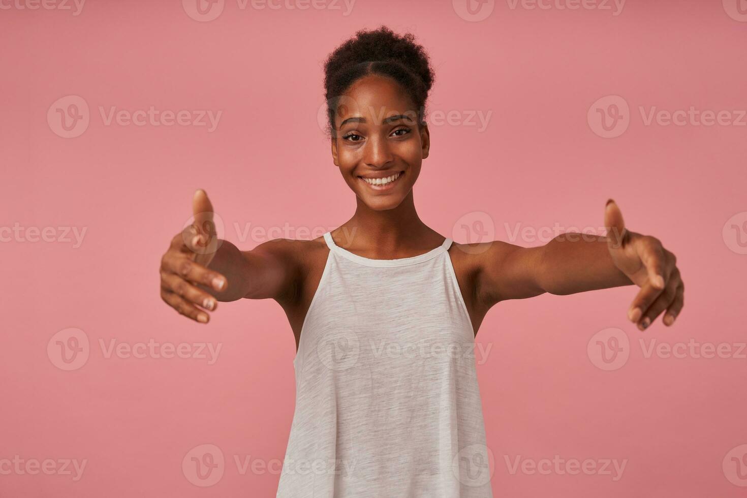 alegre jovem bonita Castanho cabelos encaracolado mulher com Sombrio pele sorridente alegremente às Câmera enquanto em pé sobre Rosa fundo com aberto braços, indo para dar abraço para alguém foto