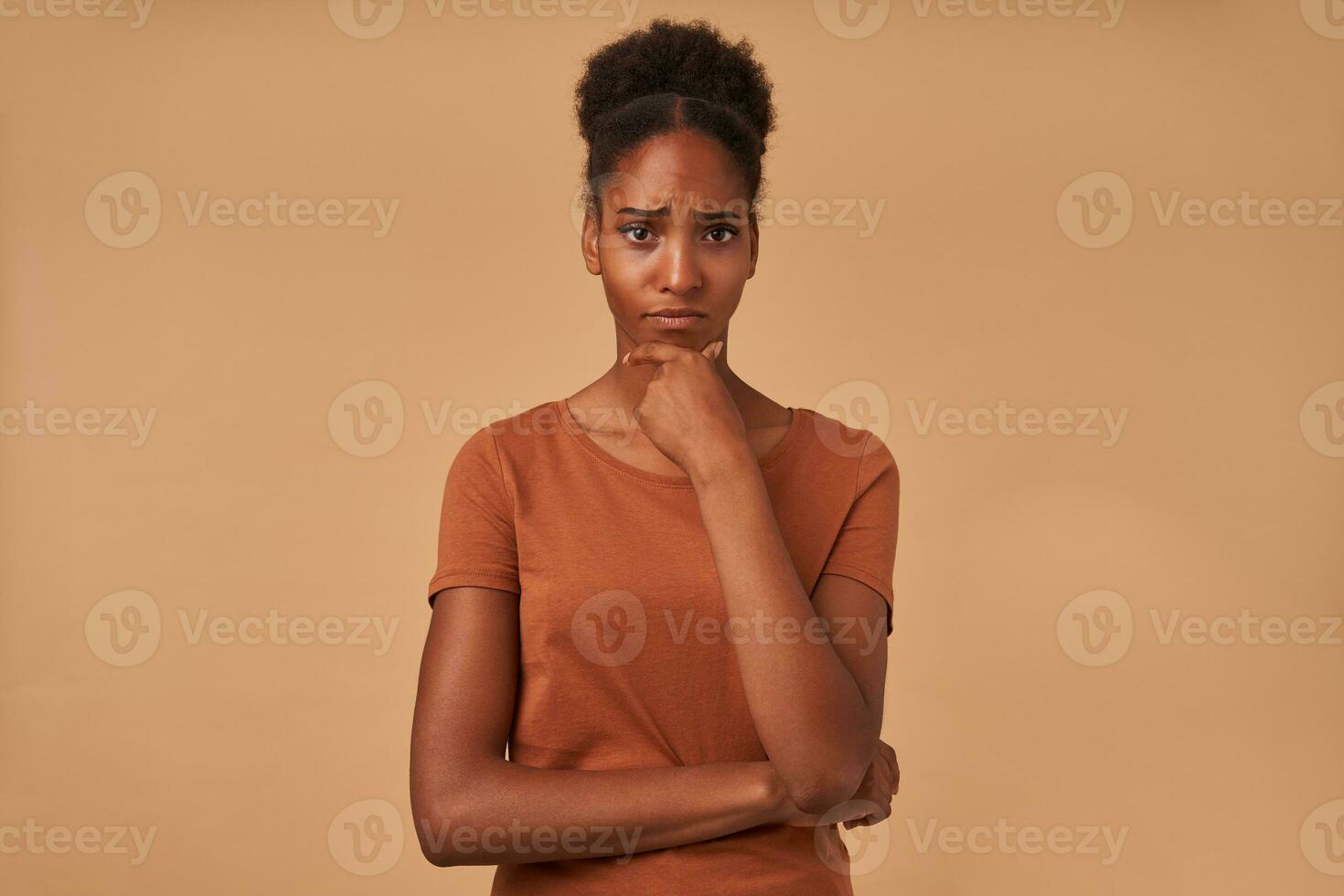 interior tiro do pensativo jovem olhos castanhos Sombrio esfolado mulher guardando elevado mão em dela queixo e olhando a sério às Câmera, posando sobre bege fundo foto