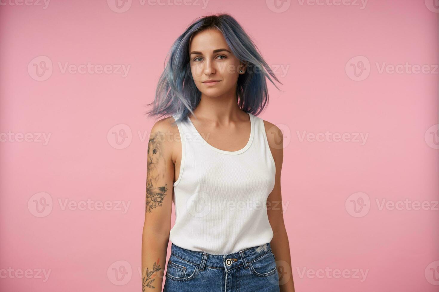 estúdio foto do atraente jovem tatuado senhora guardando mãos baixa enquanto acenando dela azul curto cabelo, vestido dentro branco camisa e jeans enquanto posando sobre Rosa fundo