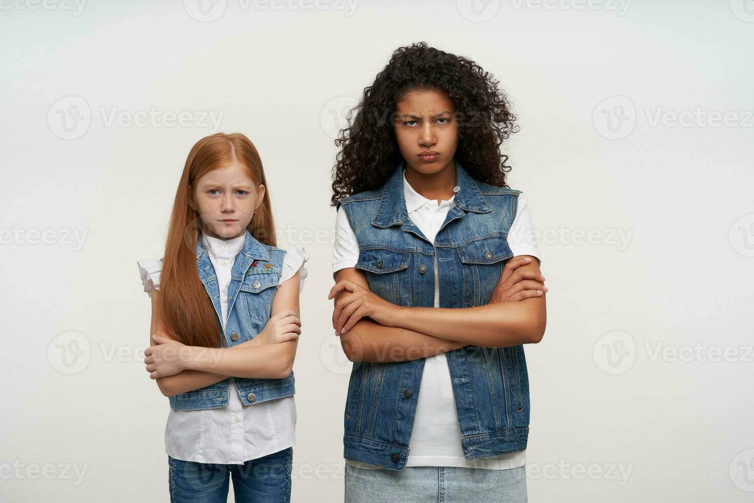 ofendido jovem Sombrio esfolado encaracolado morena senhora e vermelho cabelos bonita pequeno menina olhando Infelizmente para Câmera com guardada lábios e cruzando mãos em peito, isolado sobre branco fundo foto