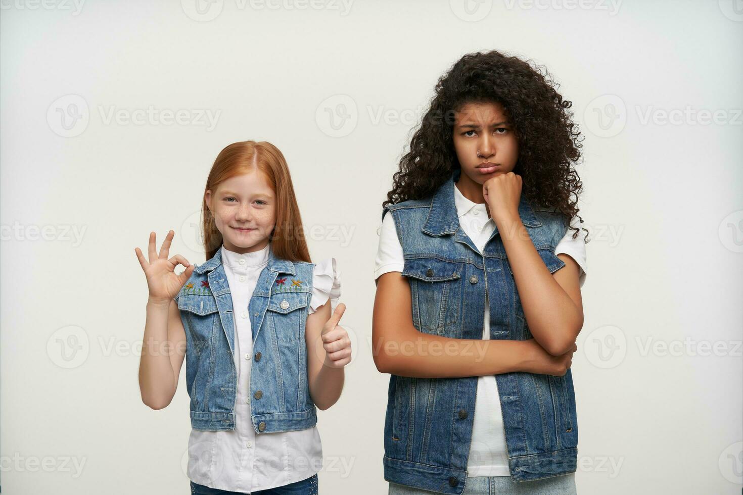 lamentoso jovem Sombrio esfolado fêmea com grandes encaracolado cabelo inclinado dela queixo em elevado punho e olhando lamentavelmente às Câmera, em pé sobre branco fundo com vermelho cabelos otimista pequeno menina foto