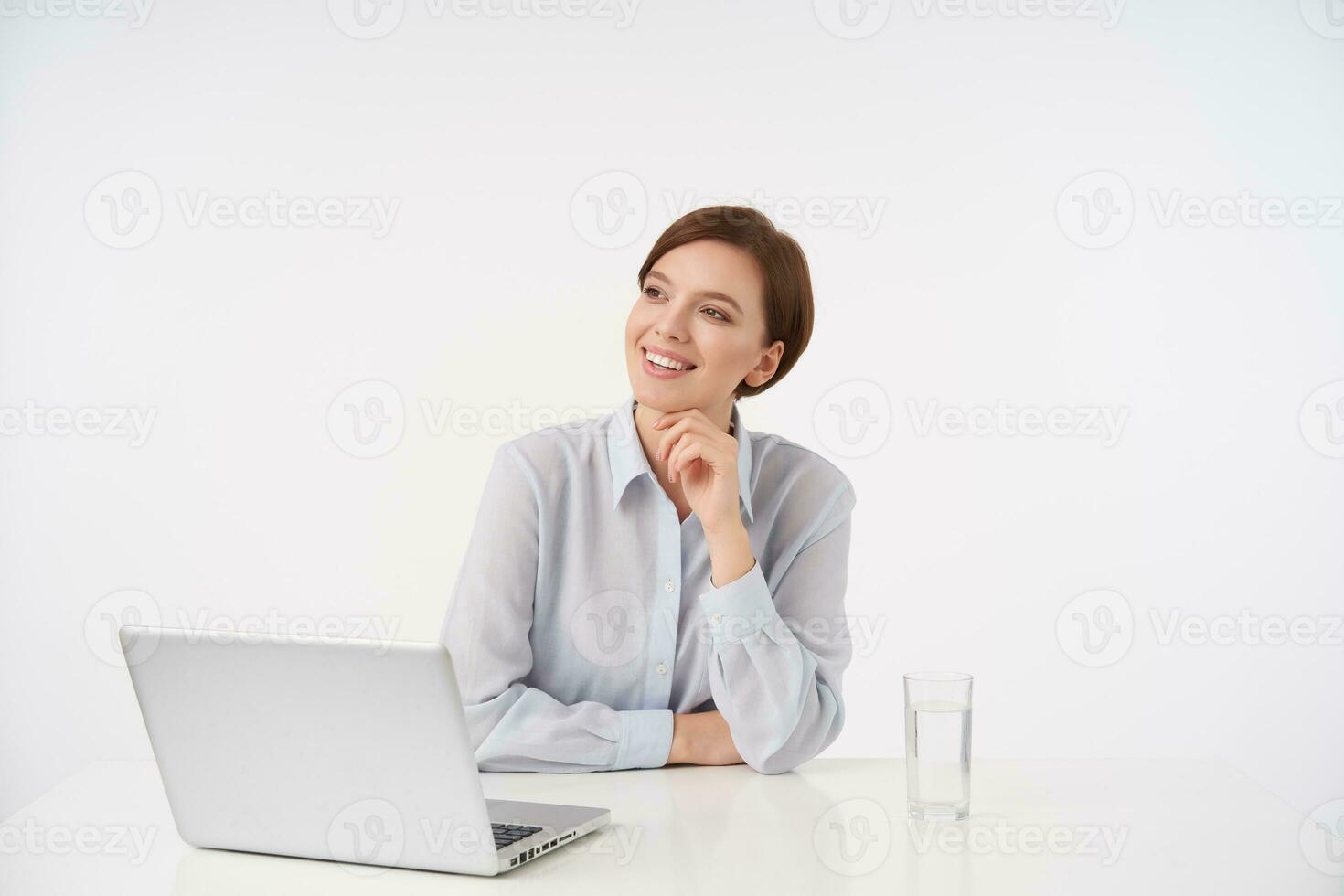 feliz jovem adorável morena mulher com casual Penteado trabalhando dentro moderno escritório com computador portátil, sorridente agradavelmente enquanto olhando a parte, de lado e segurando queixo com elevado mão, isolado sobre branco fundo foto