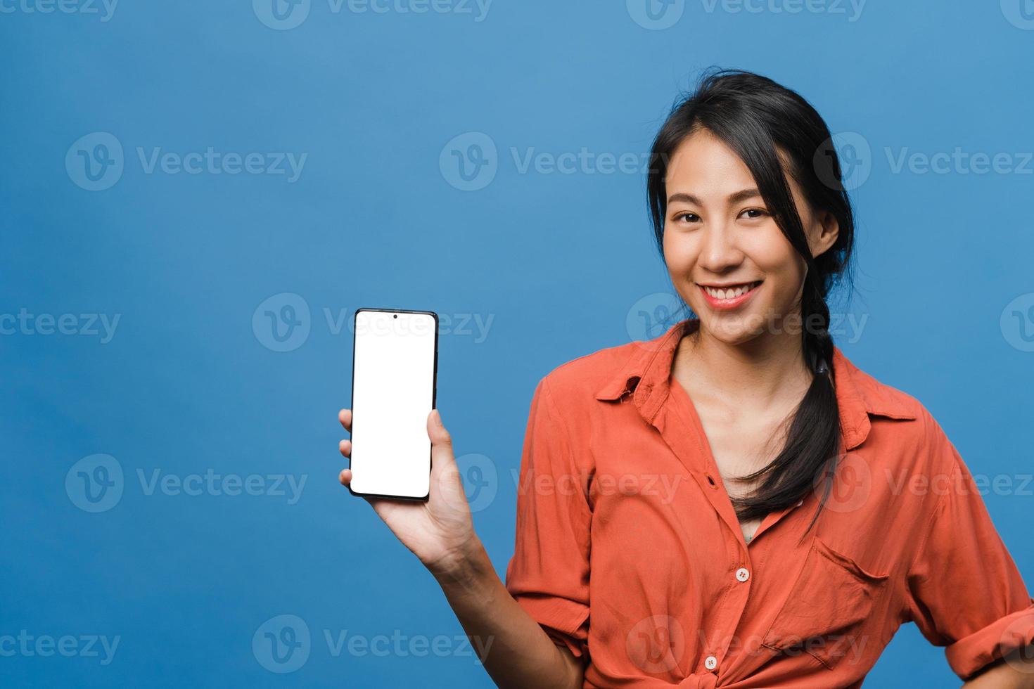 jovem asiática mostra tela vazia do smartphone com expressão positiva, sorri amplamente, vestida com roupas casuais, sentindo felicidade sobre fundo azul. telefone celular com tela branca na mão feminina. foto