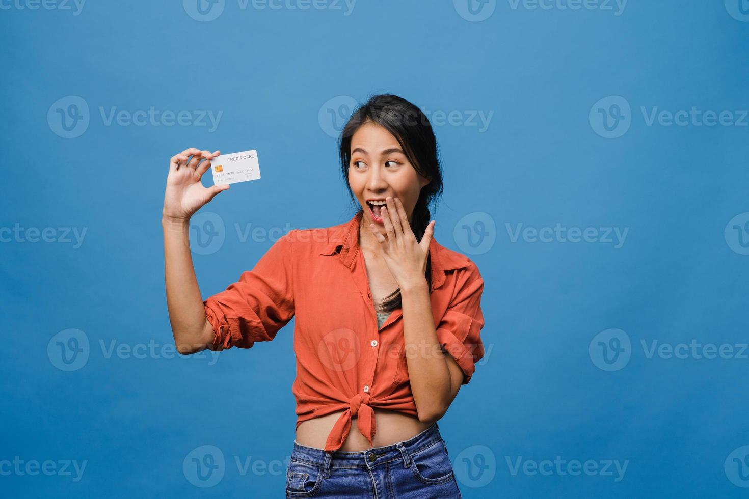 jovem senhora asiática mostra cartão de crédito com expressão positiva, sorri amplamente, vestida com roupas casuais, sentindo felicidade e carrinho isolado sobre fundo azul. conceito de expressão facial. foto