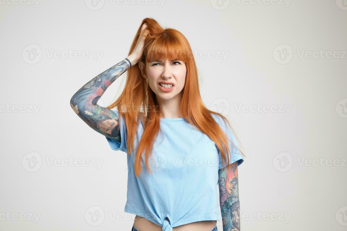 descontente jovem atraente ruiva tatuado mulher agarrando dela cabeça com elevado mão e franzindo a testa face enquanto olhando aparte, isolado sobre branco fundo foto