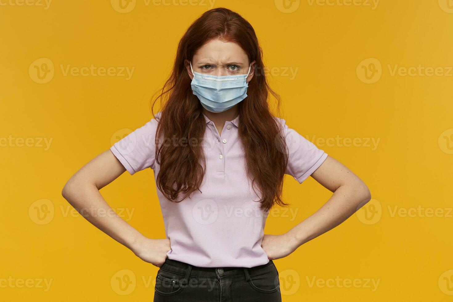 louco irritado jovem mulher vestindo médico protetora mascarar mantém mãos em cintura e olhando às Câmera sobre amarelo fundo foto