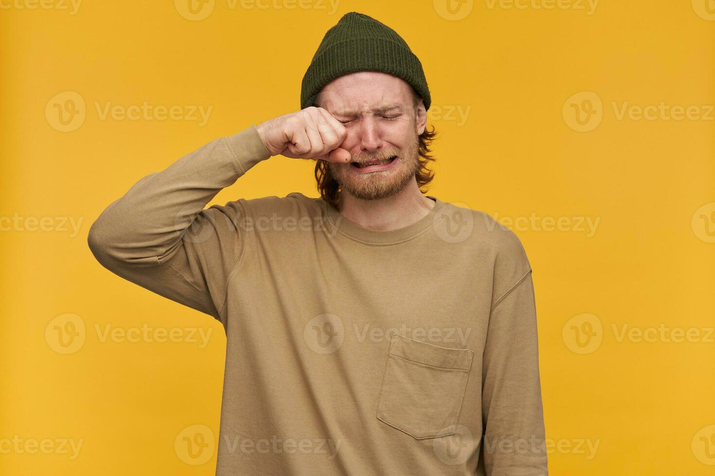 retrato do desesperado, adulto masculino com loiro cabelo e barba. vestindo verde gorro e bege suéter. chorando e lenços dele olho a partir de lágrimas. ficar de pé isolado sobre amarelo fundo foto