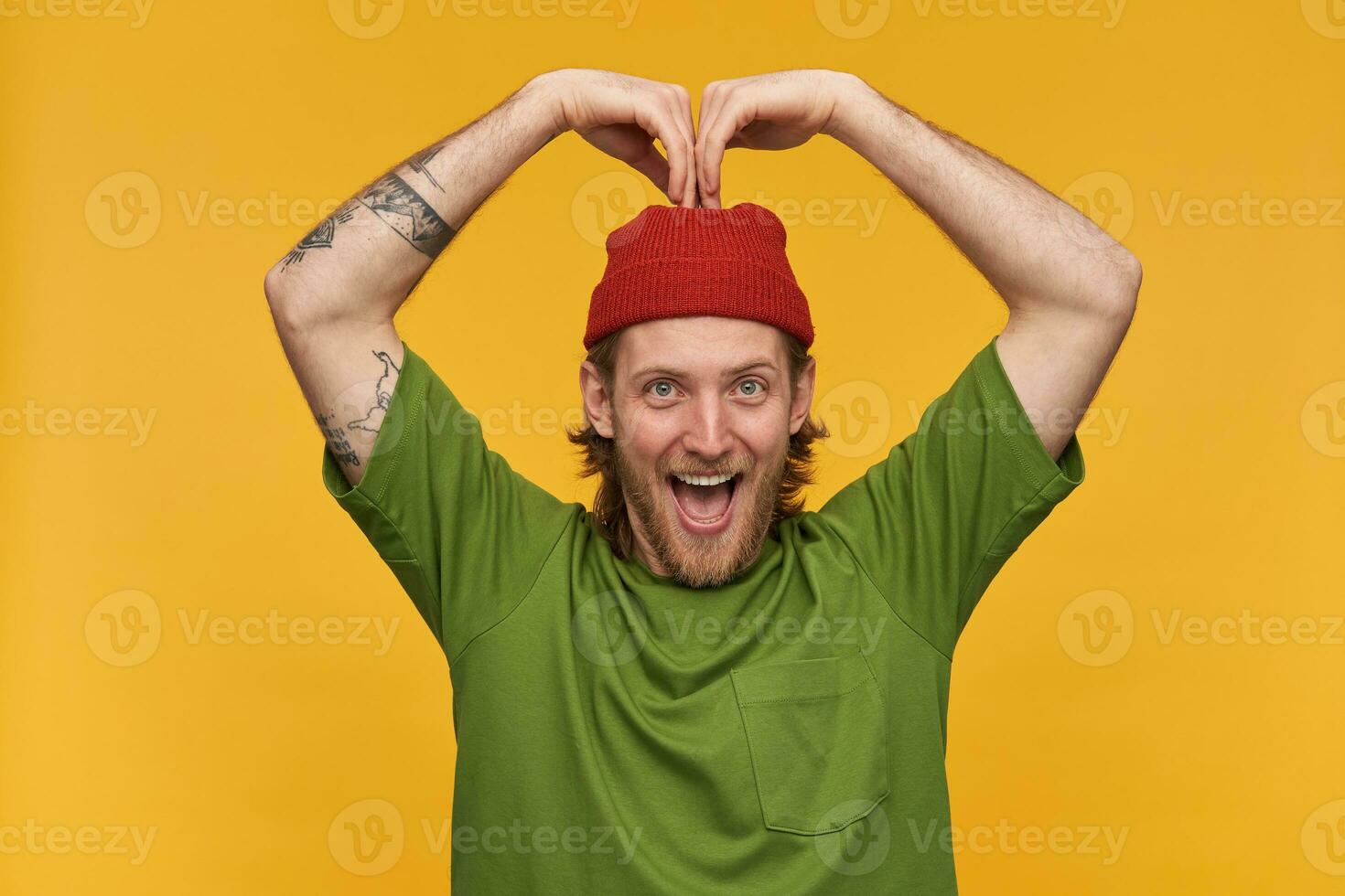 positivo, bonito barbudo cara com loiro cabelo. vestindo verde camiseta e vermelho gorro. tem tatuagens. fazer coração placa com mãos sobre dele cabeça. assistindo às a Câmera isolado sobre amarelo fundo foto