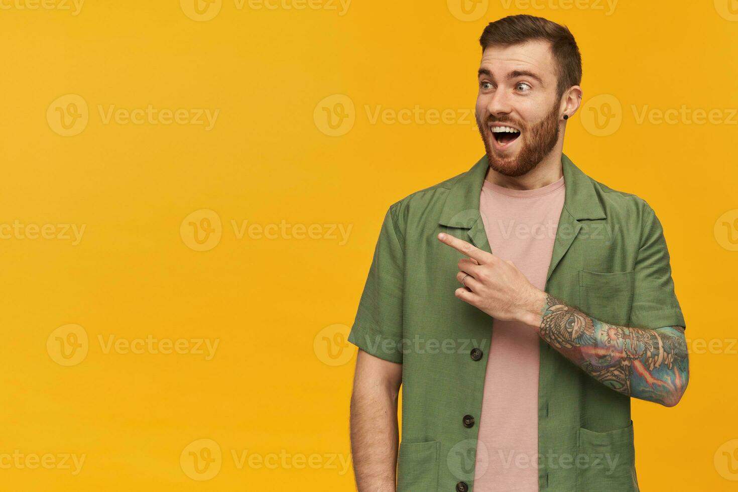 espantado feliz barbudo jovem homem com tatuagem e aberto boca olhando e apontando longe às copyspace sobre amarelo fundo foto