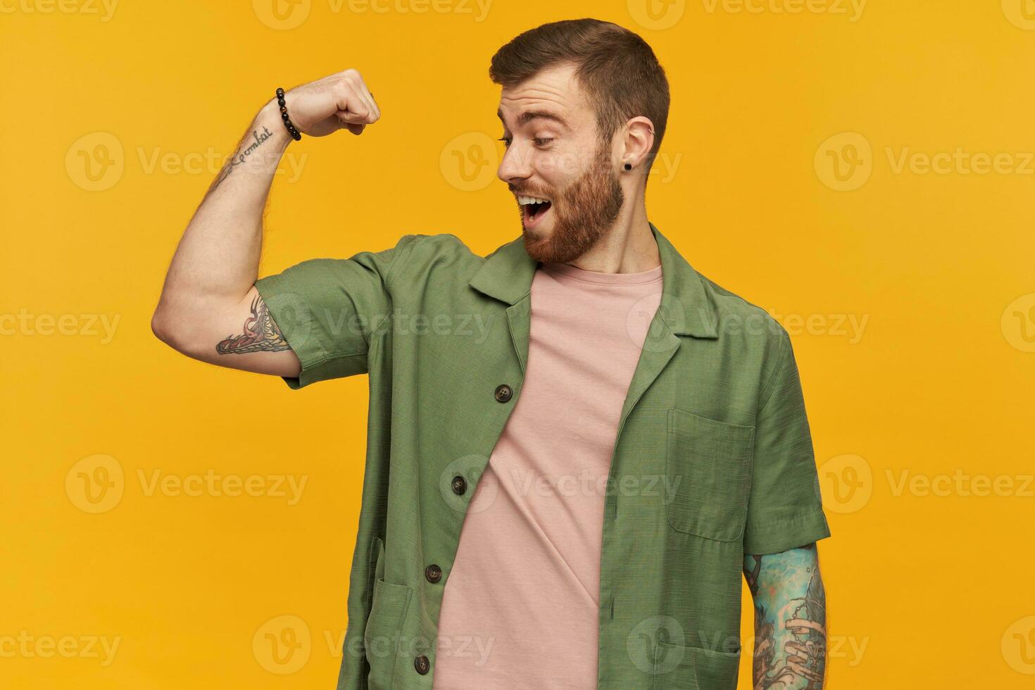 espantado animado barbudo jovem homem com tatuagem olhando e mostrando bíceps músculos sobre amarelo fundo foto