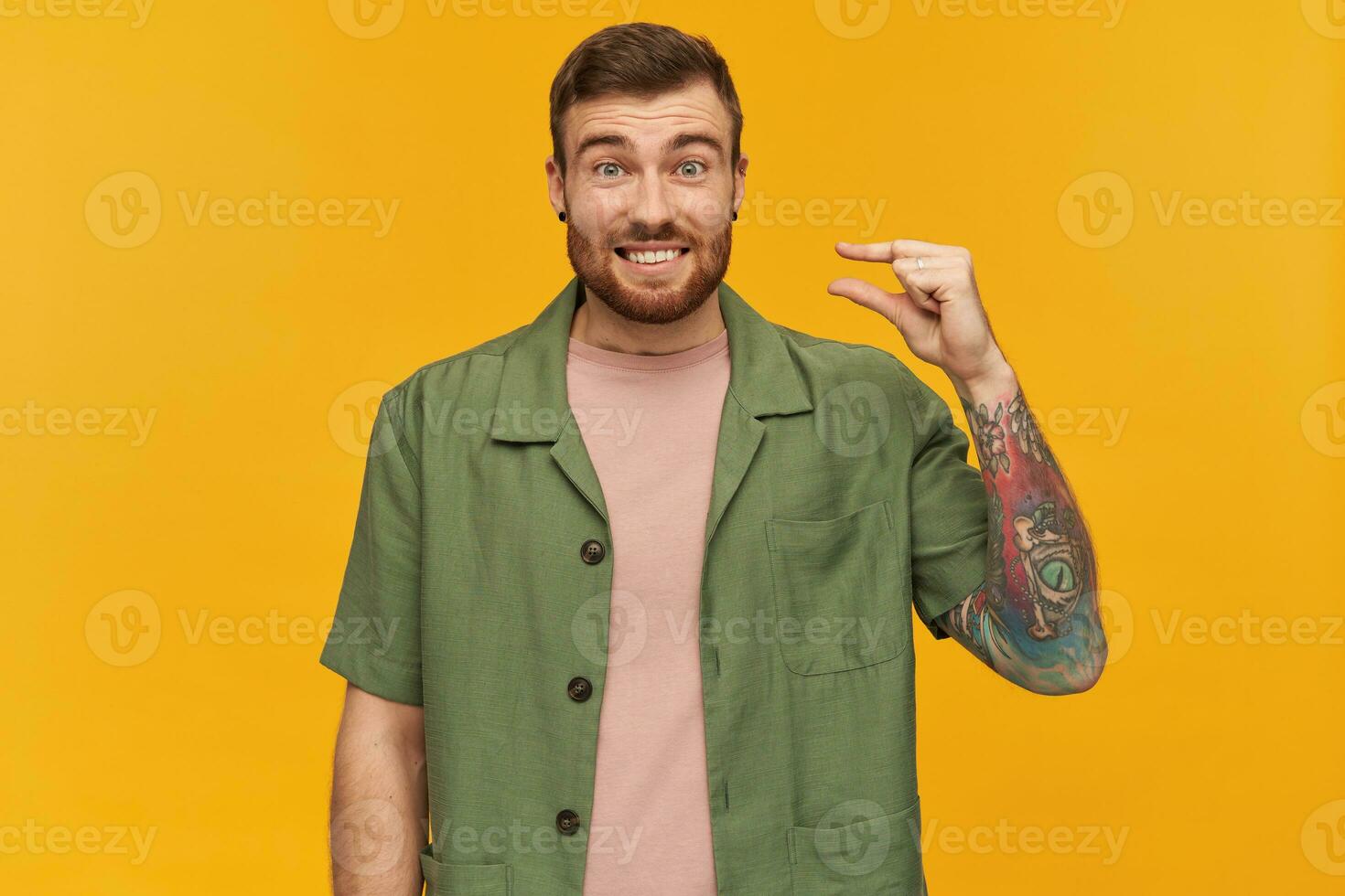 sorridente bonito jovem homem com barba e tatuagem mostrando muito pequeno Tamanho de dedos sobre amarelo fundo foto