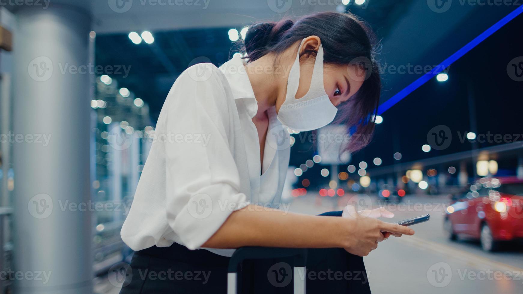 garota de negócios asiática chegar destino usar máscara facial ficar do lado de fora olhar terminal de carro de espera de telefone inteligente no aeroporto doméstico. pandemia de covid de viajante de negócios, conceito de distanciamento social de viagens de negócios. foto