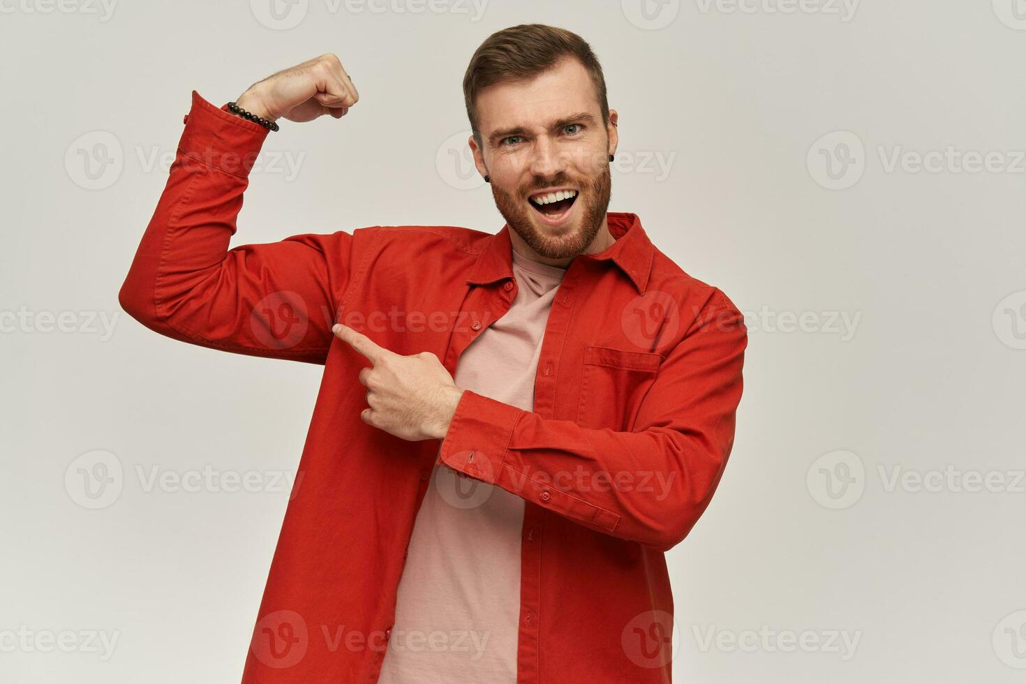 sorridente Forte jovem barbudo homem dentro vermelho camisa parece confiante e apontando às dele bíceps sobre branco fundo foto