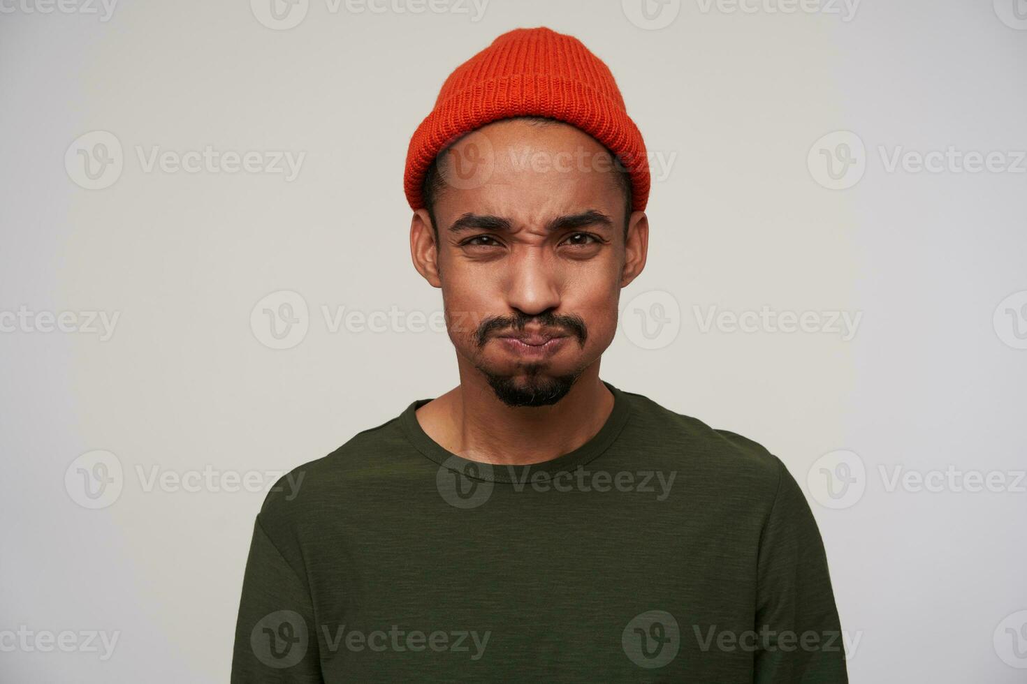 retrato do jovem olhos castanhos barbudo Sombrio esfolado masculino levando ar para dentro dele boca e bufando Fora bochechas enquanto olhando para Câmera, em pé contra branco fundo foto